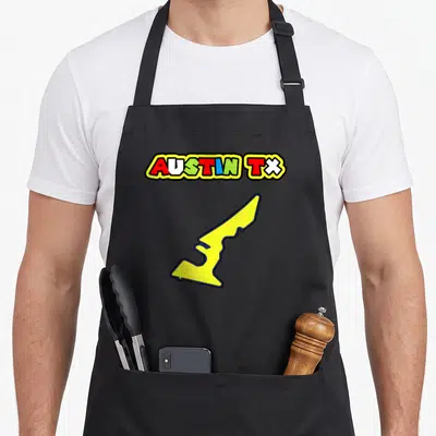 Bike texas aprons - AUSTIN TX text above yellow racetrack shape in red green blue and white cartoon style — shown on a black cotton canvas apron against a white background