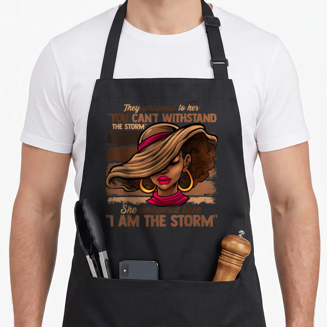 Melanin birthday diva apron - Cartoon woman in wide hat with text They whispered to her You Can't Withstand The Storm — shown on a black cotton canvas apron against a white background