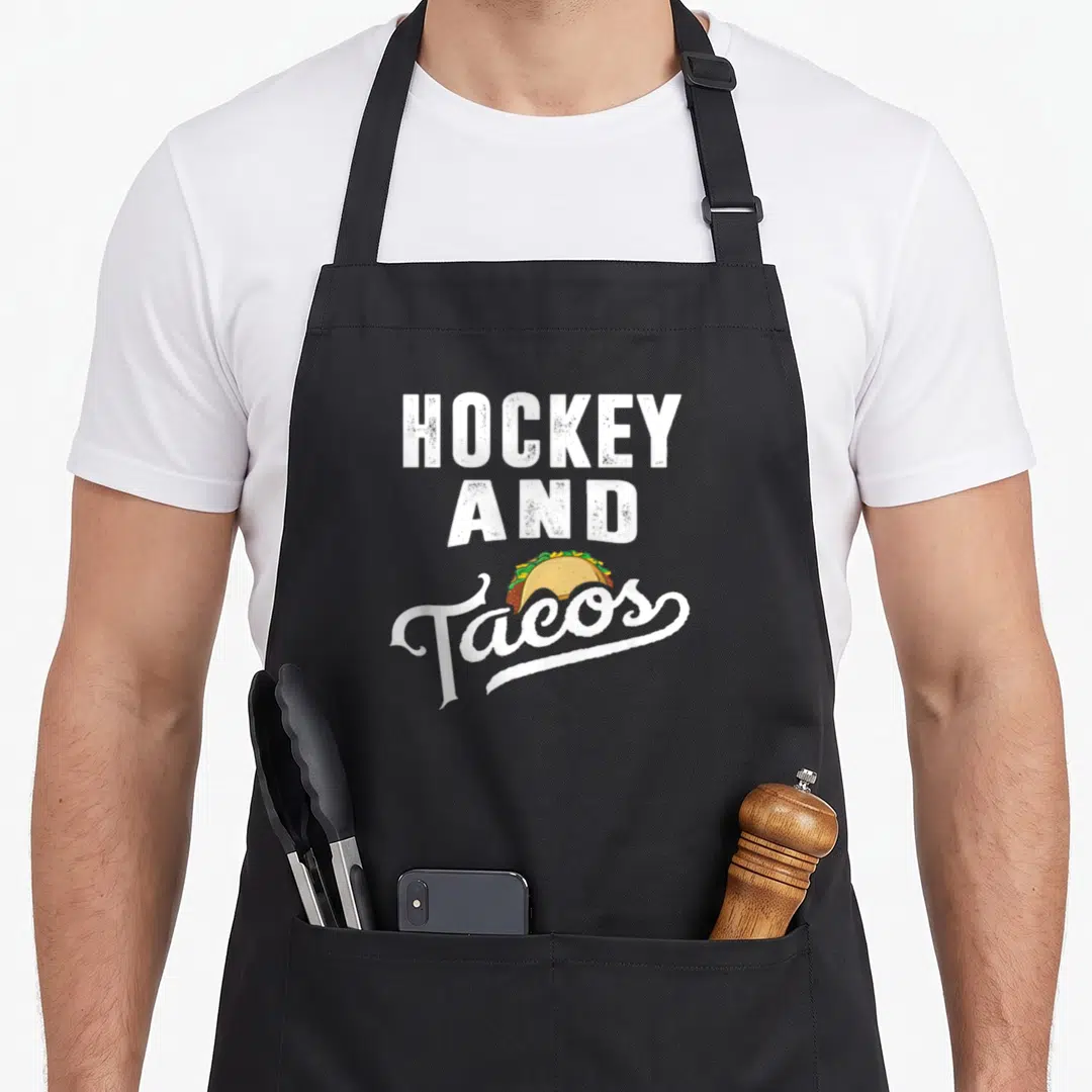 Hockey taco apron - Distressed white HOCKEY AND Tacos text with a yellow taco illustration — shown on a black cotton canvas apron against a white background