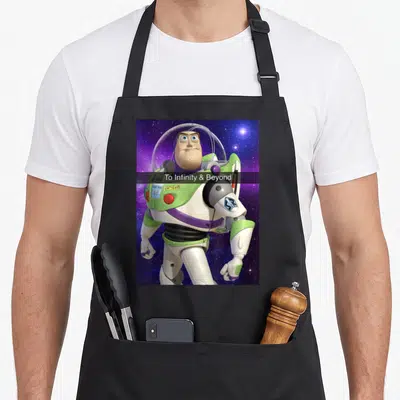 Funny beyond infinity apron - Buzz Lightyear centered on starry background with To Infinity and Beyond text on black bar — shown on a black cotton canvas apron against a white background