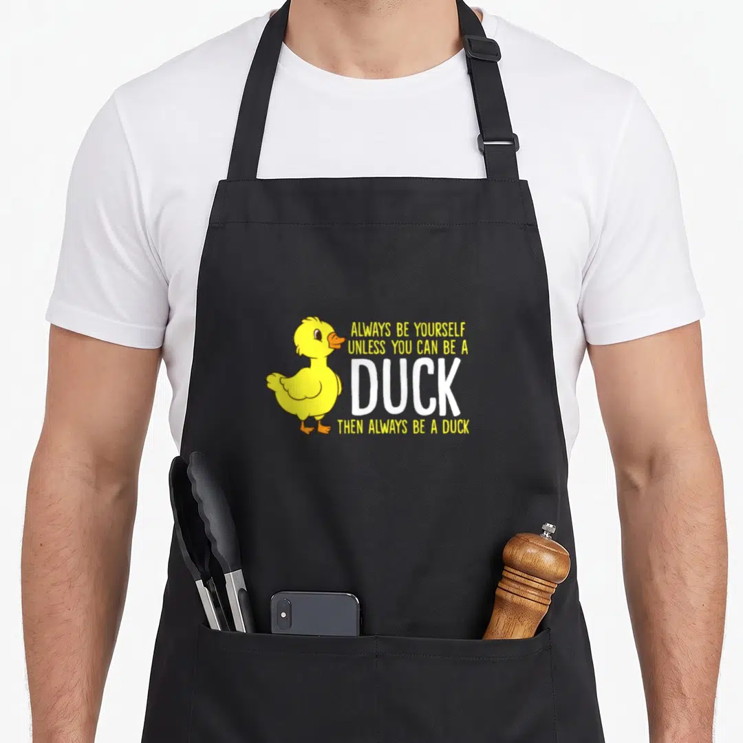 Unless duck apron - Cartoon yellow duck standing beside stacked text Always Be Yourself Unless You Can Be A Duck — shown on a black cotton canvas apron against a white background