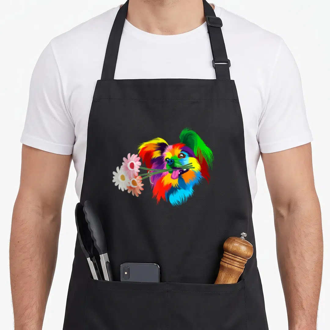 Colored dog apron - Rainbow cartoon papillon head holding three daisies in mouth — shown on a black cotton canvas apron against a white background