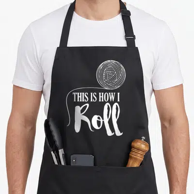 Roll craft apron - White yarn ball illustration with text THIS IS HOW I Roll in mixed typography — shown on a black cotton canvas apron against a white background