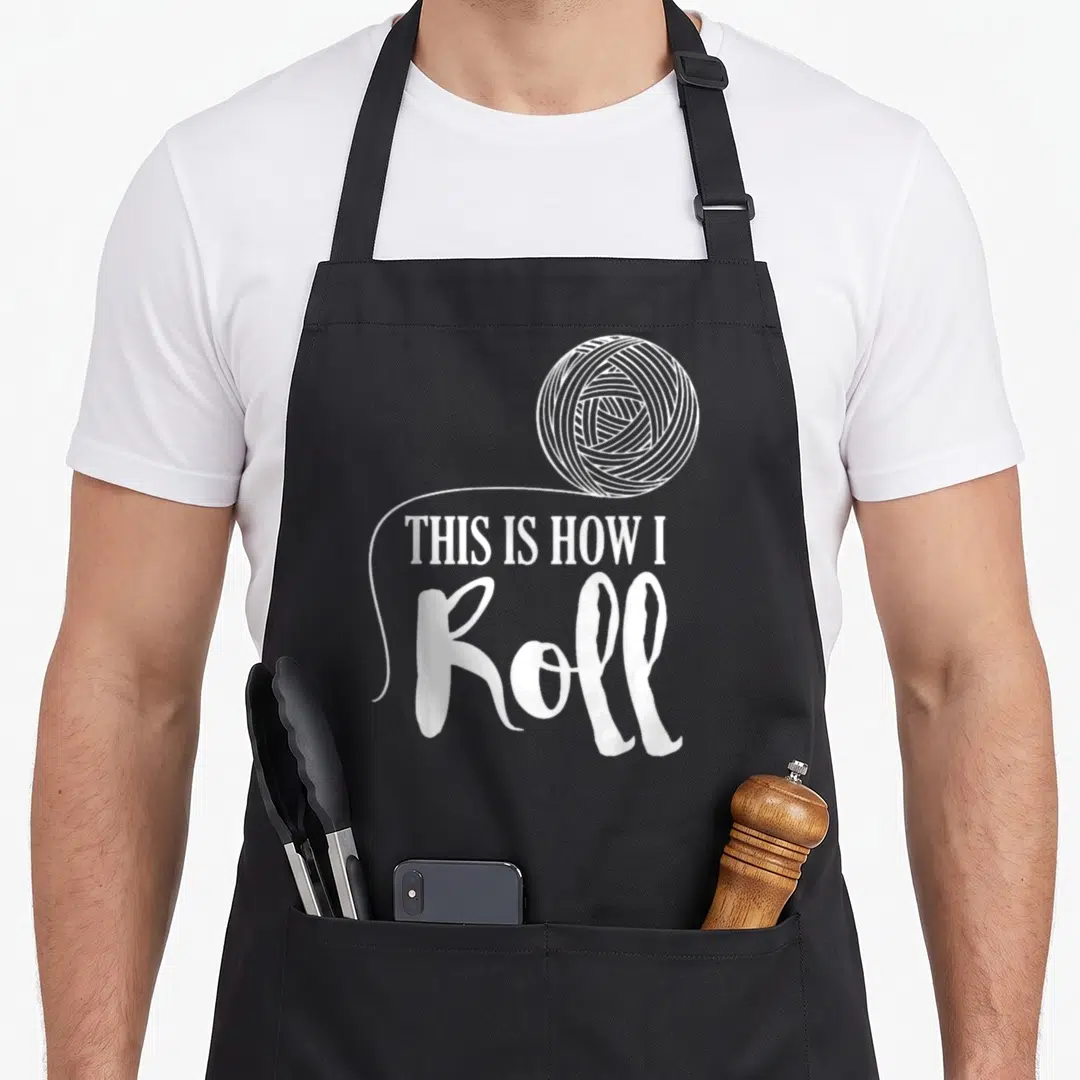 Roll craft apron - White yarn ball illustration with text THIS IS HOW I Roll in mixed typography — shown on a black cotton canvas apron against a white background