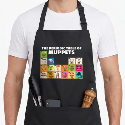Funny periodic table apron - Muppet character heads in colored grid squares with symbols Kf Kermit Mp Miss Piggy Gz — shown on a black cotton canvas apron against a white background