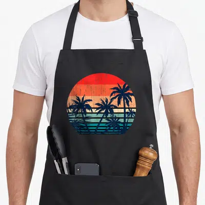 Summer vacation tree apron - Retro palm tree silhouettes against a circular sun with red orange and blue horizontal — shown on a black cotton canvas apron against a white background