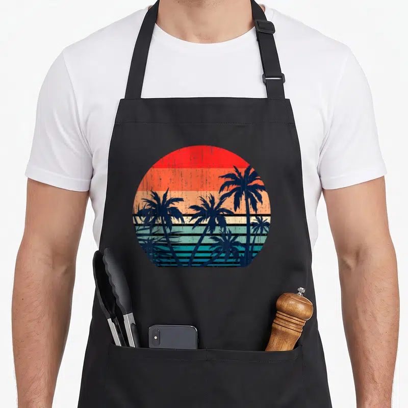 Summer vacation tree apron - Retro palm tree silhouettes against a circular sun with red orange and blue horizontal — shown on a black cotton canvas apron against a white background