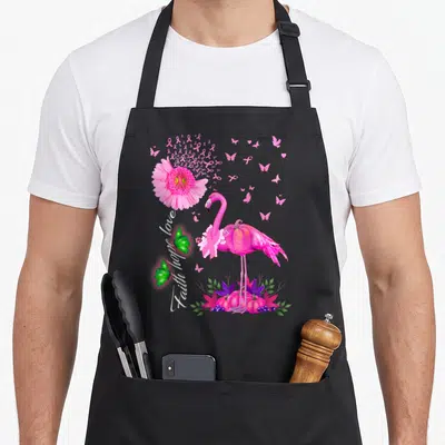 Faith pink flamingo apron - Pink flamingo wearing ribbon stands on pink pumpkins beside vertical Faith Hope Love text — shown on a black cotton canvas apron against a white background