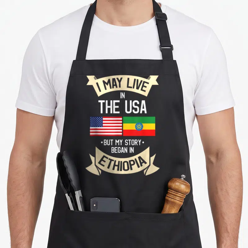 Ethiopian ethiopia apron - Stacked text with USA and Ethiopia flags side by side in beige ribbons using white red blue — shown on a black cotton canvas apron against a white background