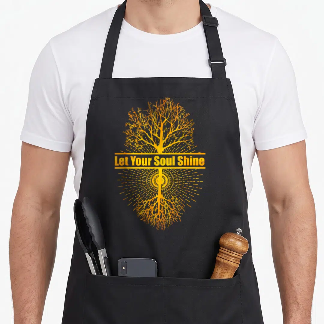 Shine tree apron - Orange and yellow stylized tree with roots and Let Your Soul Shine text over sunburst — shown on a black cotton canvas apron against a white background