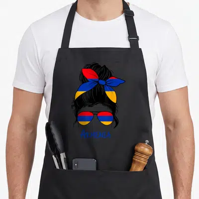 Flag armenian apron - Cartoon woman head with bun and sunglasses filled with red blue orange stripes, ARMENIA text below — shown on a black cotton canvas apron against a white background