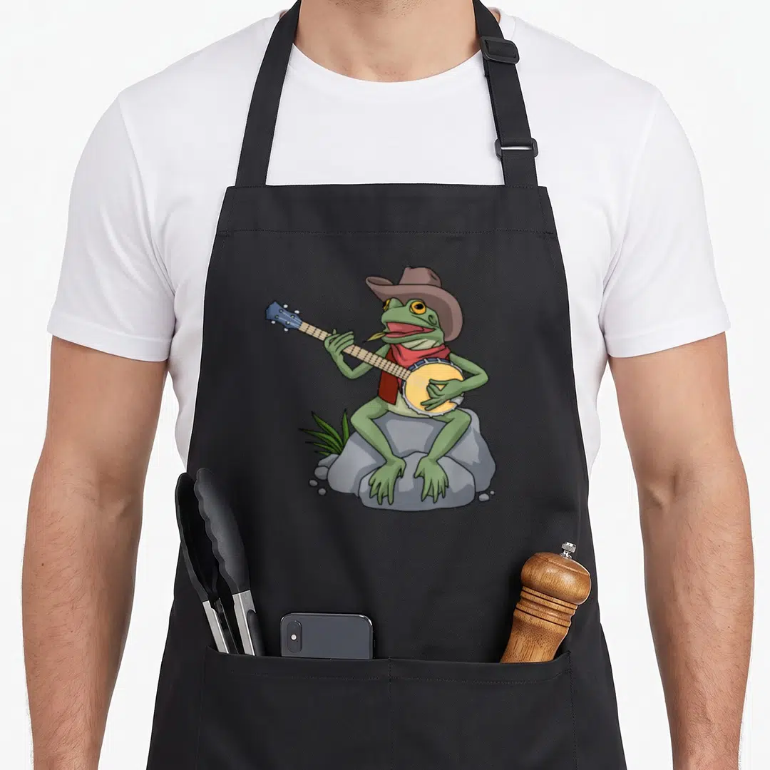 Bluegrass cowboy apron - Cartoon frog in brown hat and red bandana playing banjo on grey rock — shown on a black cotton canvas apron against a white background