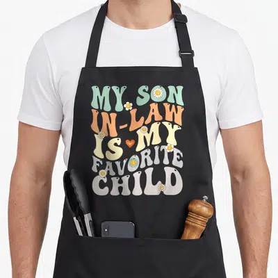 Law son humor apron - Retro bubbly text reading My Son In Law Is My Favorite Child with smiley daisies and a heart — shown on a black cotton canvas apron against a white background
