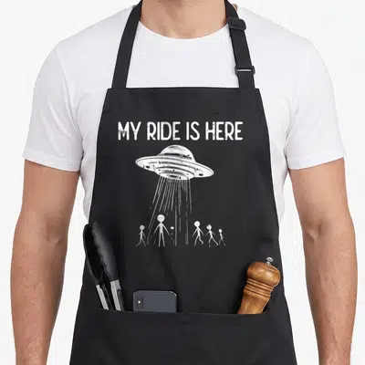 Alien flying apron - White UFO and five stick figures under MY RIDE IS HERE text — shown on a black cotton canvas apron against a white background