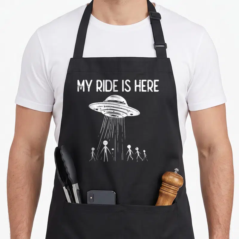 Alien flying apron - White UFO and five stick figures under MY RIDE IS HERE text — shown on a black cotton canvas apron against a white background