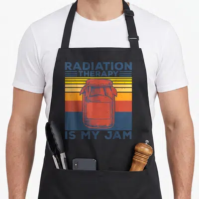 Costume radiation aprons - Retro red jam jar over blue yellow orange stripes with RADIATION THERAPY IS MY JAM text — shown on a black cotton canvas apron against a white background