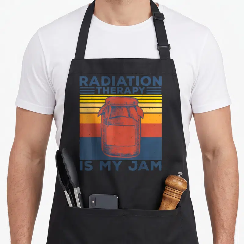 Costume radiation aprons - Retro red jam jar over blue yellow orange stripes with RADIATION THERAPY IS MY JAM text — shown on a black cotton canvas apron against a white background