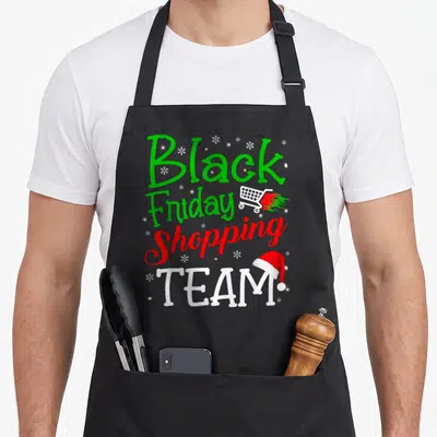 Shopping christmas black friday apron - Stacked green red white text Black Friday Shopping TEAM with flaming cart — shown on a black cotton canvas apron against a white background