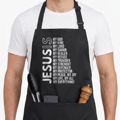 Christianity christian apron - Vertical JESUS IS text beside stacked list of divine titles in distressed white font — shown on a black cotton canvas apron against a white background