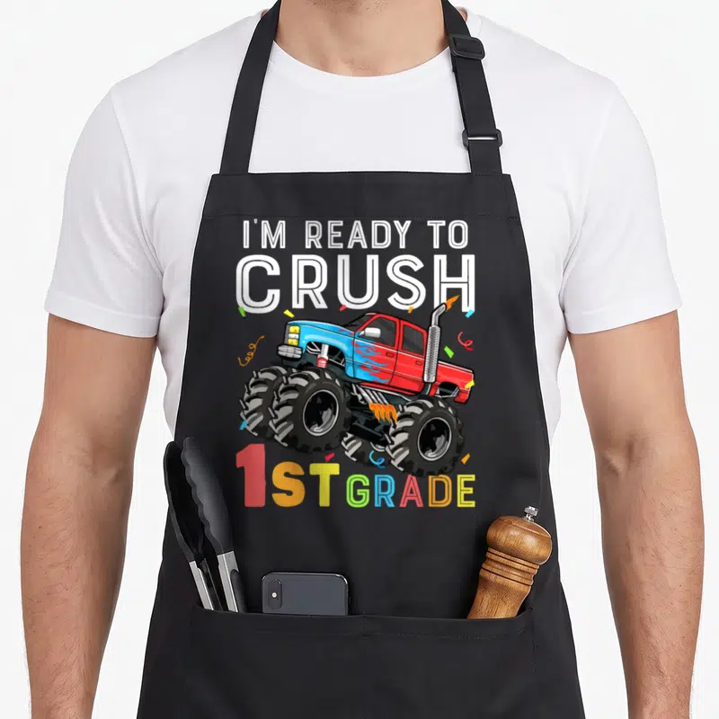 Ready grade apron - I'M READY TO CRUSH 1ST GRADE monster truck with red blue yellow green confetti — shown on a black cotton canvas apron against a white background