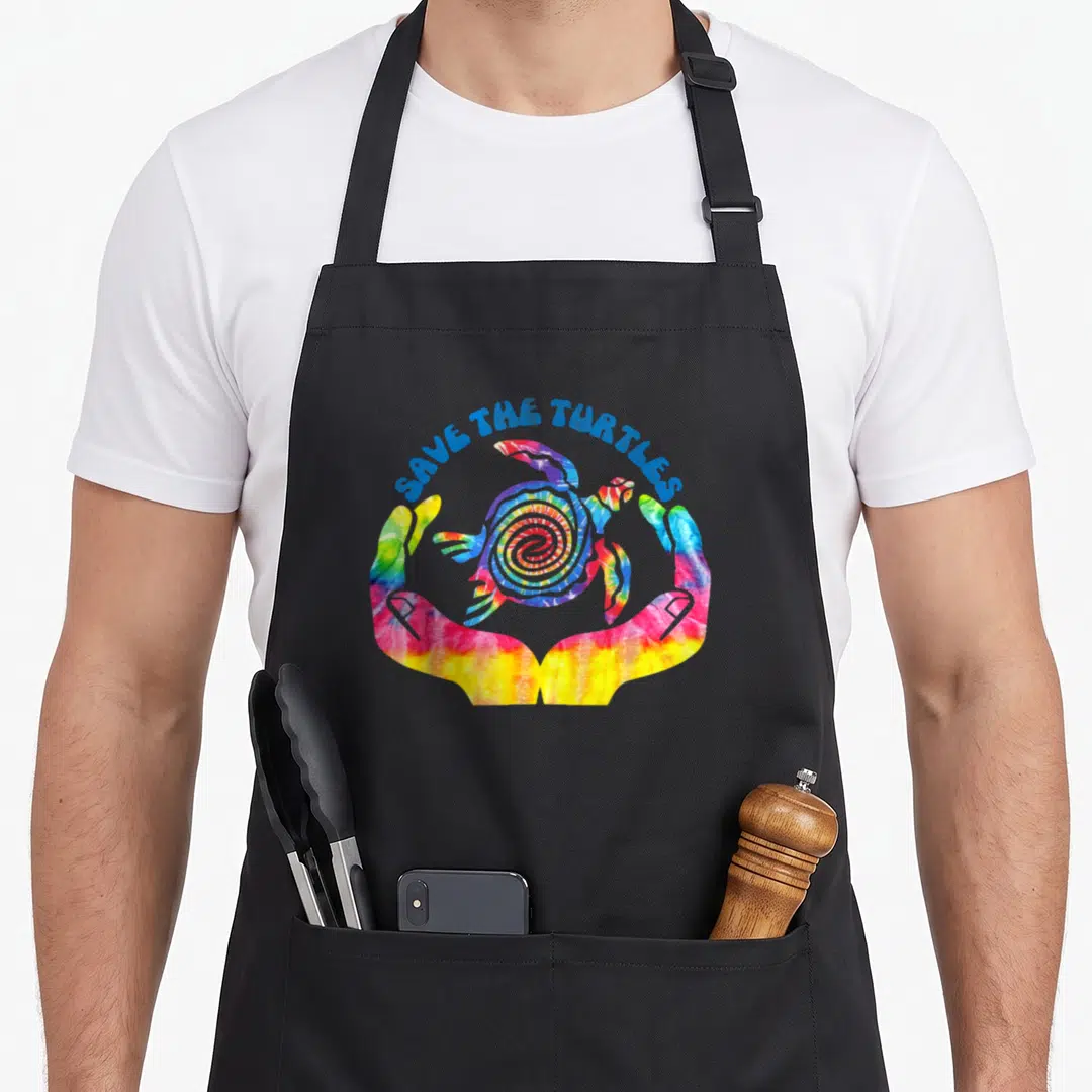 Sea conservation apron - Tie-dye turtle cradled by hands with blue arched text SAVE THE TURTLES above — shown on a black cotton canvas apron against a white background