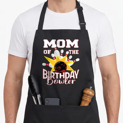 Bowler birthday apron - MOM OF THE BIRTHDAY Bowler text surrounding a black bowling ball hitting pins with a yellow — shown on a black cotton canvas apron against a white background