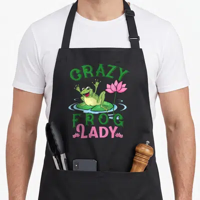 Frog grandma's aprons - Cartoon frog on lily pad with CRAZY FROG LADY text and pink lotus flower — shown on a black cotton canvas apron against a white background