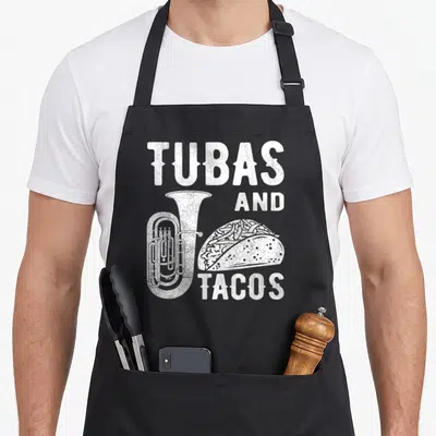 Tuba taco apron - White distressed TUBAS AND TACOS text with tuba and taco illustrations — shown on a black cotton canvas apron against a white background