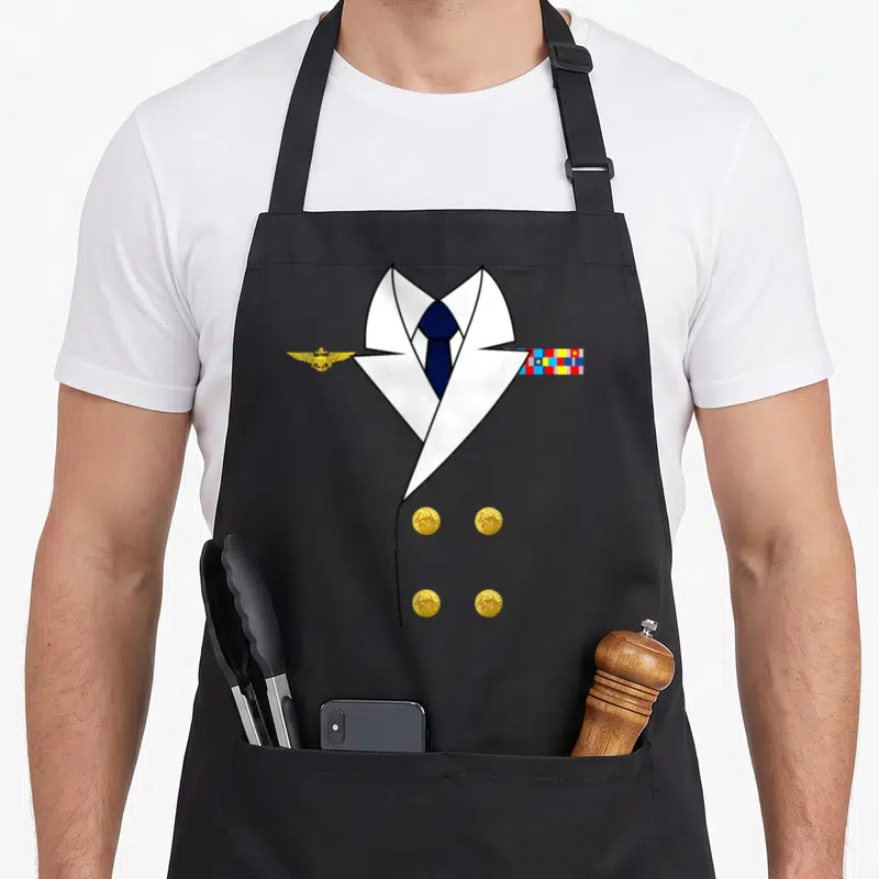 Ship halloween apron - White uniform jacket, blue tie, gold wings, multicolored ribbons, four gold buttons — shown on a black cotton canvas apron against a white background