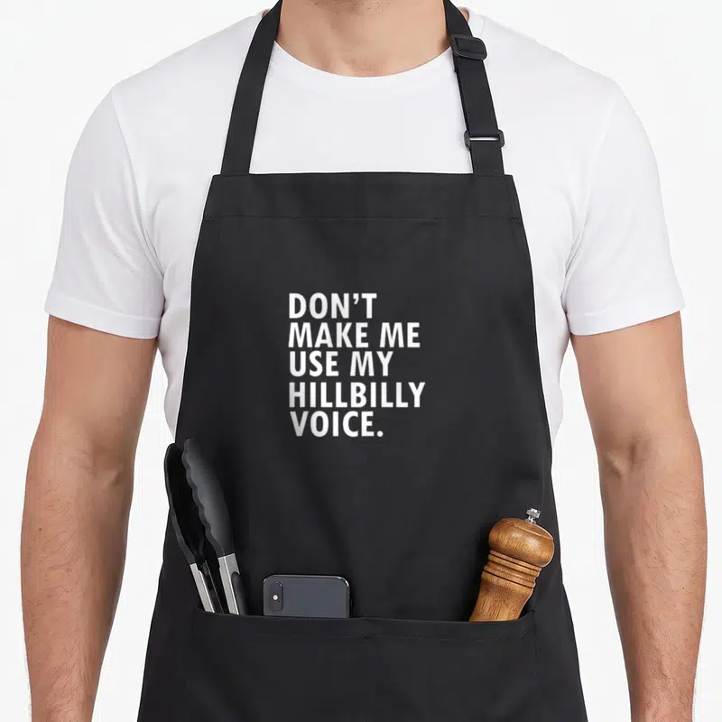 Hillbilly southern aprons - White stacked text reading DON'T MAKE ME USE MY HILLBILLY VOICE on grey background — shown on a black cotton canvas apron against a white background