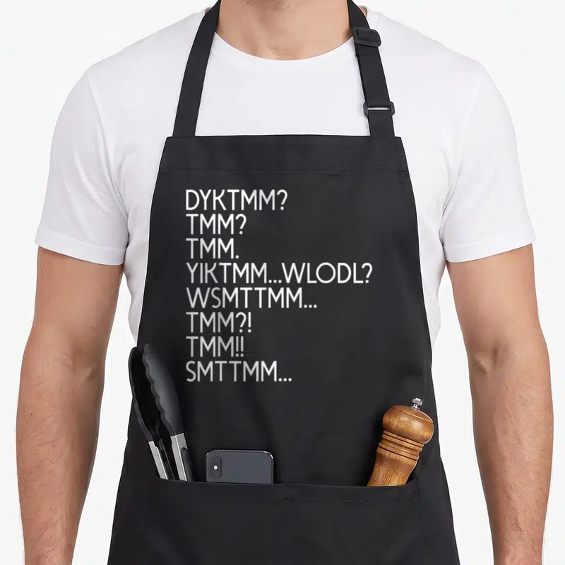 Funny dyktmm muffin man apron - Eight lines of white text including DYKTMM and TMM abbreviations stacked vertically — shown on a black cotton canvas apron against a white background