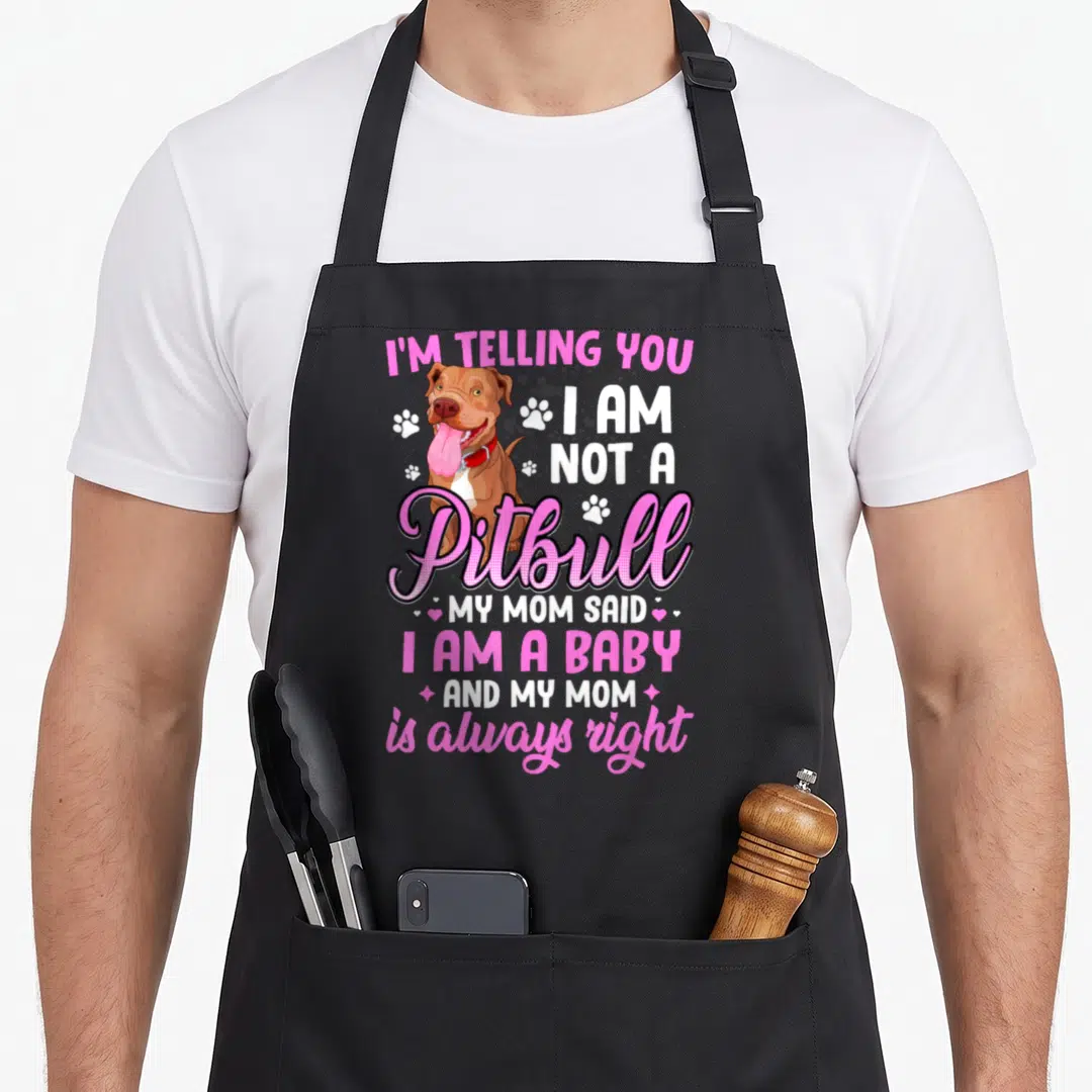 Telling pitbull apron - Cartoon brown dog with pink text reading I Am Not A Pitbull My Mom Said I Am A Baby — shown on a black cotton canvas apron against a white background