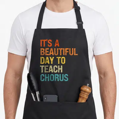 Chorus music teacher apron - Stacked retro text reading It's a Beautiful Day to Teach Chorus in orange yellow beige — shown on a black cotton canvas apron against a white background