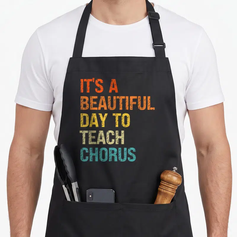 Chorus music teacher apron - Stacked retro text reading It's a Beautiful Day to Teach Chorus in orange yellow beige — shown on a black cotton canvas apron against a white background
