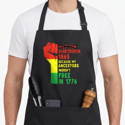 Ancestor african apron - Raised fist in red yellow green bands next to crossed out July 4th and Juneteenth 1865 text — shown on a black cotton canvas apron against a white background