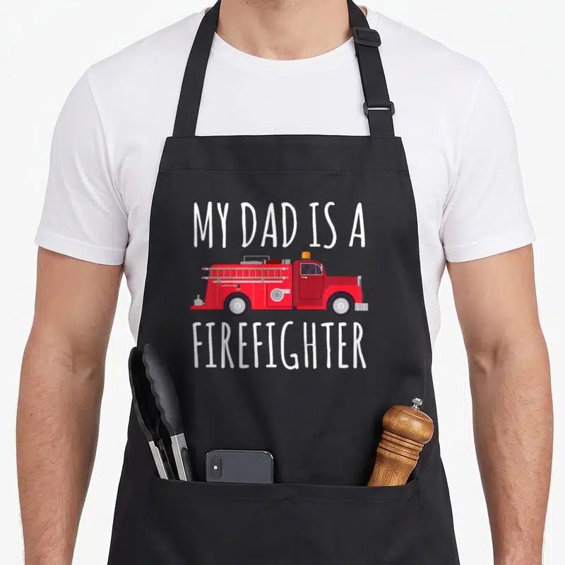 Funny firefighter aprons - Red fire truck between white MY DAD IS A FIREFIGHTER text on grey background — shown on a black cotton canvas apron against a white background