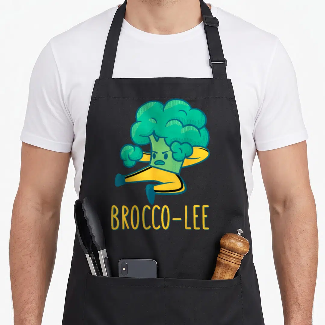 Game vegetable apron - Cartoon broccoli kicking in yellow suit with text BROCCO-LEE — shown on a black cotton canvas apron against a white background