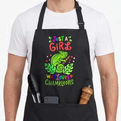 Raptile chameleon apron - Cartoon green chameleon on branch with text Just a Girl Who loves CHAMELEONS and colorful dots — shown on a black cotton canvas apron against a white background