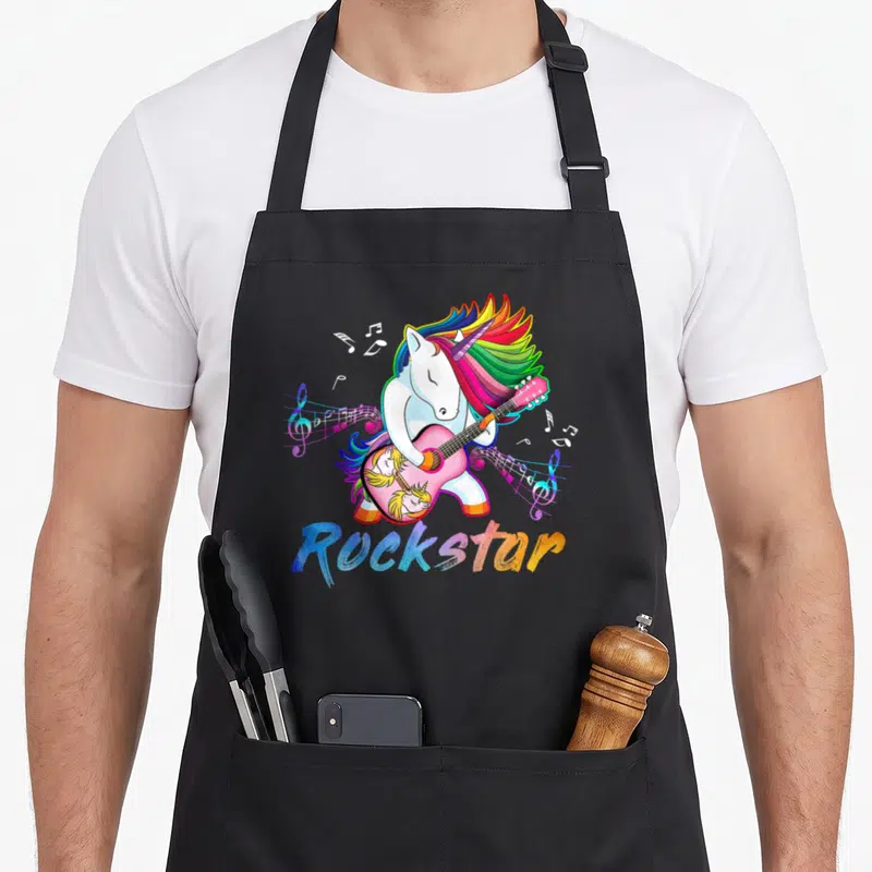 Star unicorn apron - Cartoon unicorn playing pink guitar with rainbow mane and Rockstar text — shown on a black cotton canvas apron against a white background