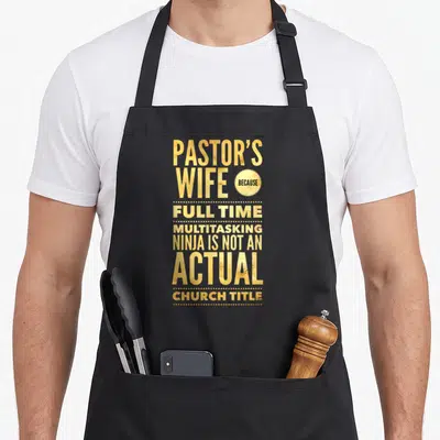Pastoral wife apron - Gold stacked text reading Pastor's Wife Because Full Time Multitasking Ninja on black background — shown on a black cotton canvas apron against a white background