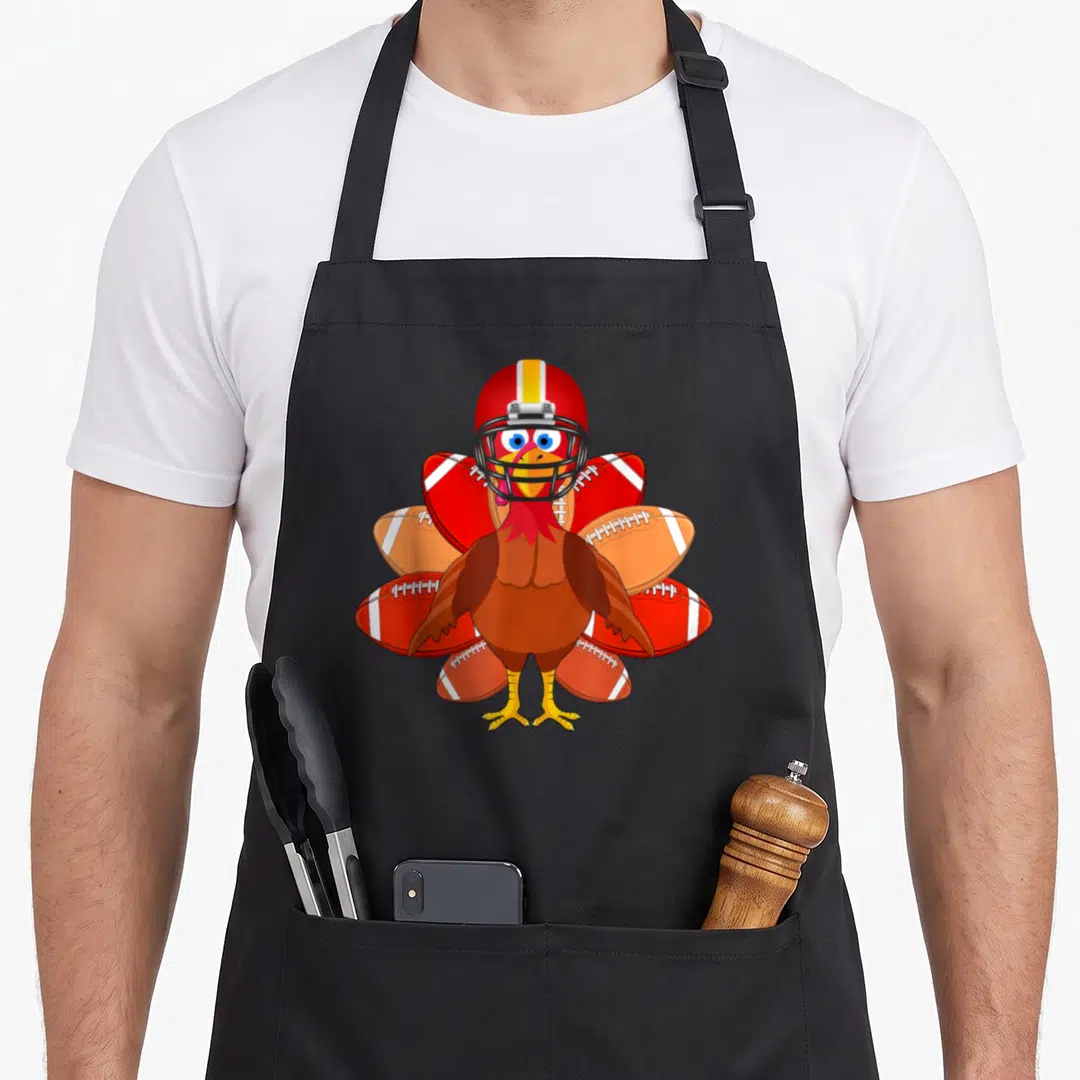 Football thankful apron - Cartoon turkey wearing a red football helmet with footballs fanned out as tail feathers — shown on a black cotton canvas apron against a white background