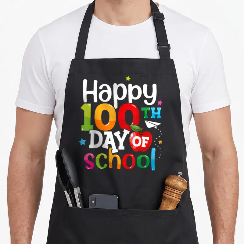 School 100th teacher apron - Stacked Happy 100th Day of School text with red apple, paper plane, and stars in white red — shown on a black cotton canvas apron against a white background