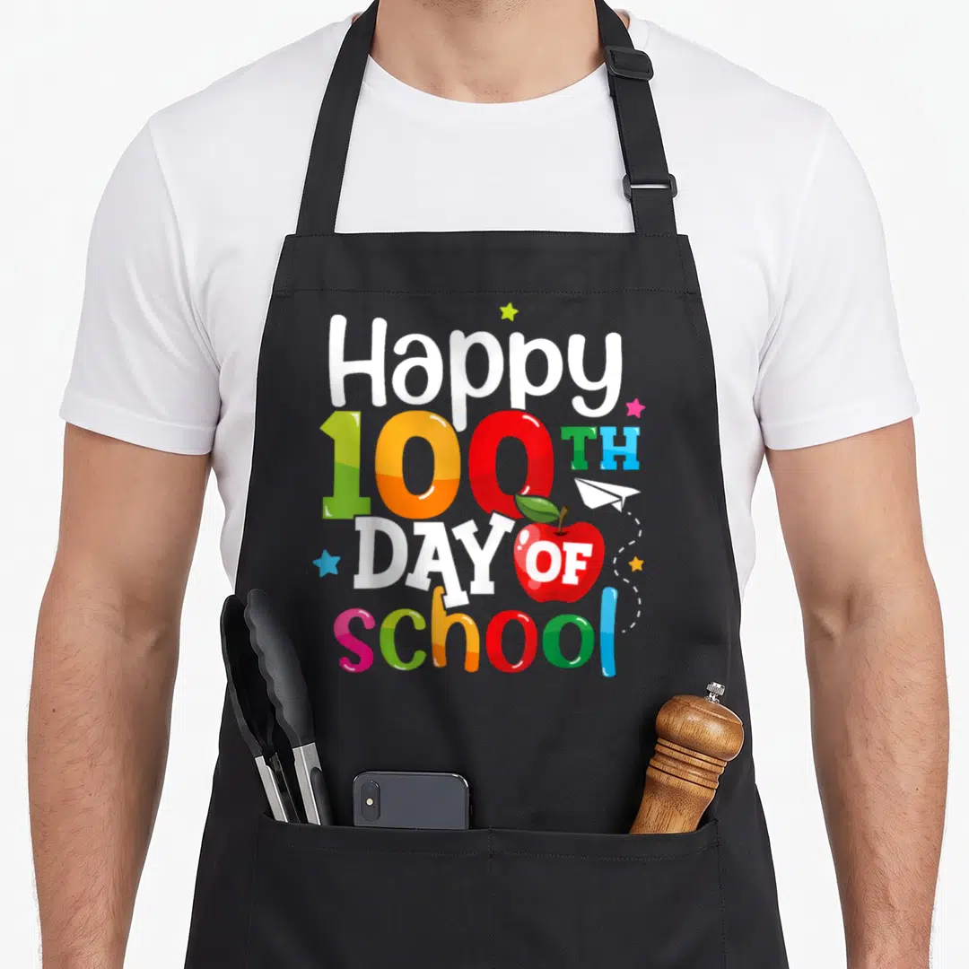 School 100th teacher apron - Stacked Happy 100th Day of School text with red apple, paper plane, and stars in white red — shown on a black cotton canvas apron against a white background