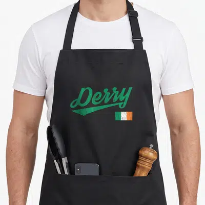Derry ireland apron - Green Derry script with swoosh and small distressed Irish flag — shown on a black cotton canvas apron against a white background