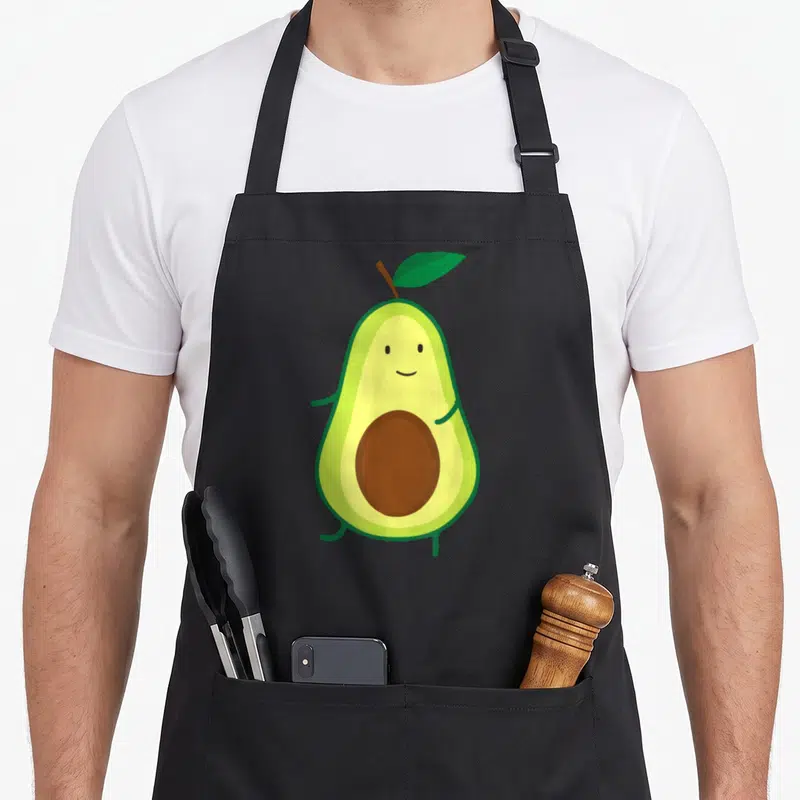 Mexican avocado apron - Smiling cartoon avocado with brown pit and green leaf — shown on a black cotton canvas apron against a white background