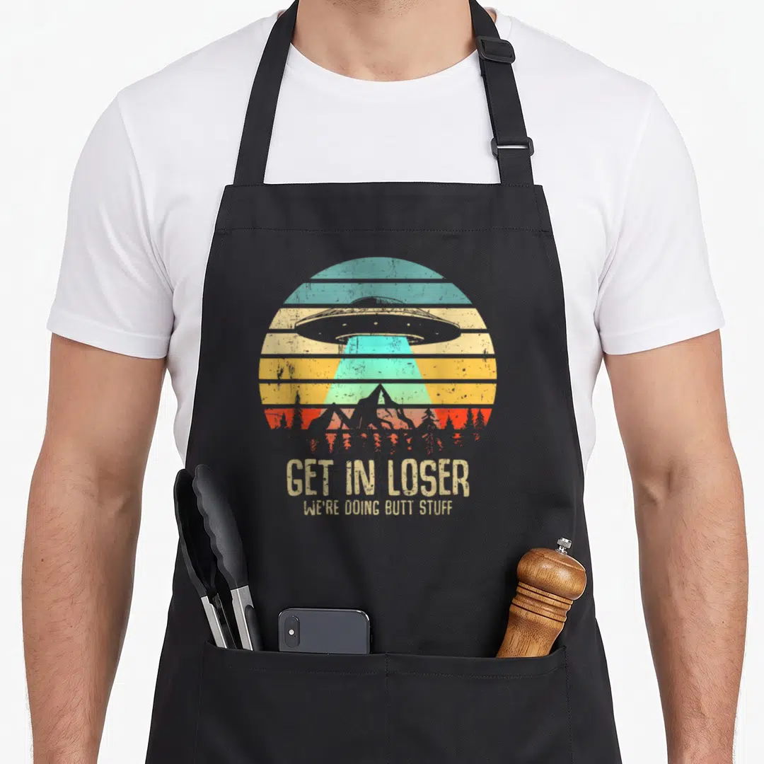 Loser skunk apron - Retro UFO over mountains with text GET IN LOSER WE'RE DOING BUTT STUFF in beige teal yellow orange — shown on a black cotton canvas apron against a white background