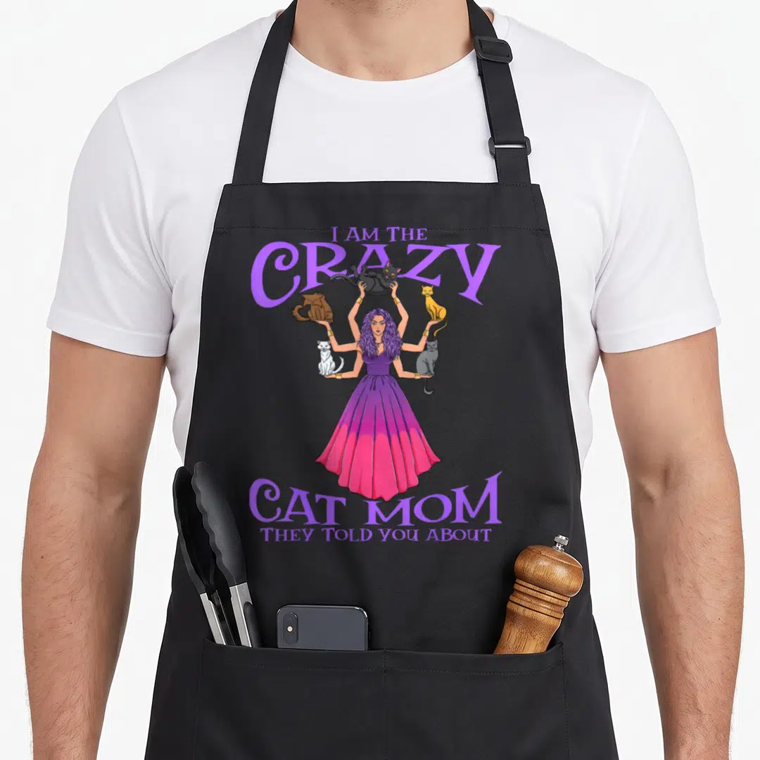 They cat apron - Woman with six arms holding five cats, purple dress, stacked text I Am The Crazy Cat Mom — shown on a black cotton canvas apron against a white background