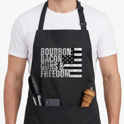 Bourbon bacon apron - Stacked BOURBON BACON GUNS FREEDOM text next to a distressed black and white flag graphic — shown on a black cotton canvas apron against a white background