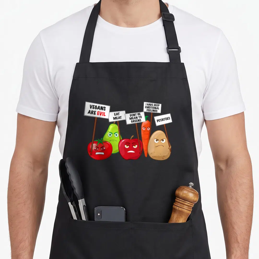 Vegan vegetable apron - Angry tomato pear apple carrot potato holding signs reading VEGANS ARE EVIL EAT MEAT — shown on a black cotton canvas apron against a white background
