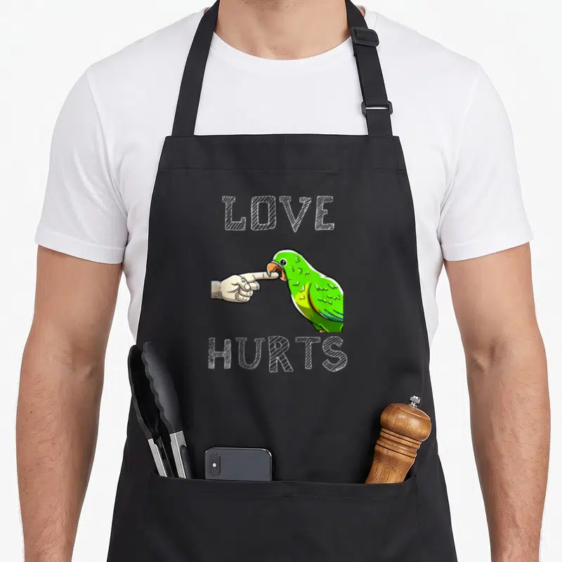Boredkoalas bird apron - Green parrot biting beige finger with LOVE HURTS text in white sketch font — shown on a black cotton canvas apron against a white background